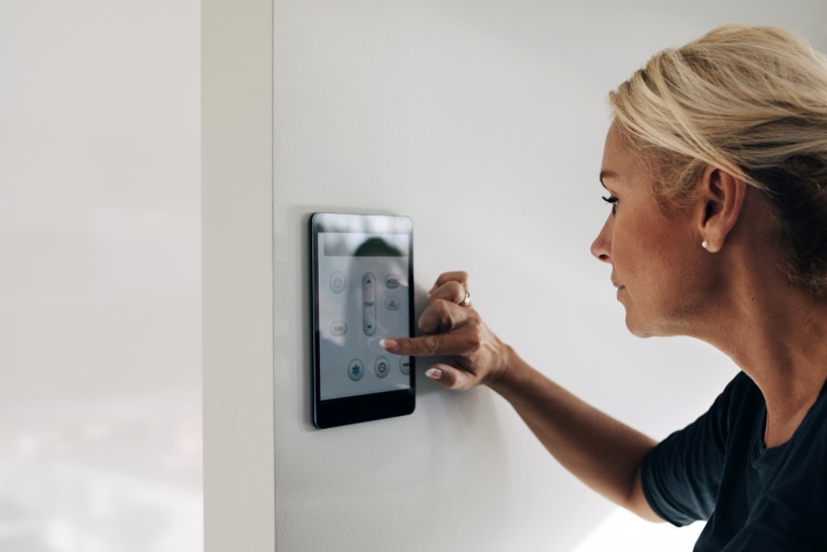 Image: Blond woman adjusting thermostat using digital tablet mounted on white wall at home